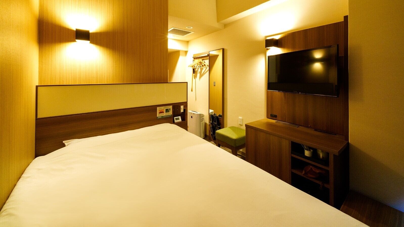 A hotel room with a bed, a flat-screen TV mounted on the wall, a small green stool, and a wooden cabinet below the TV.