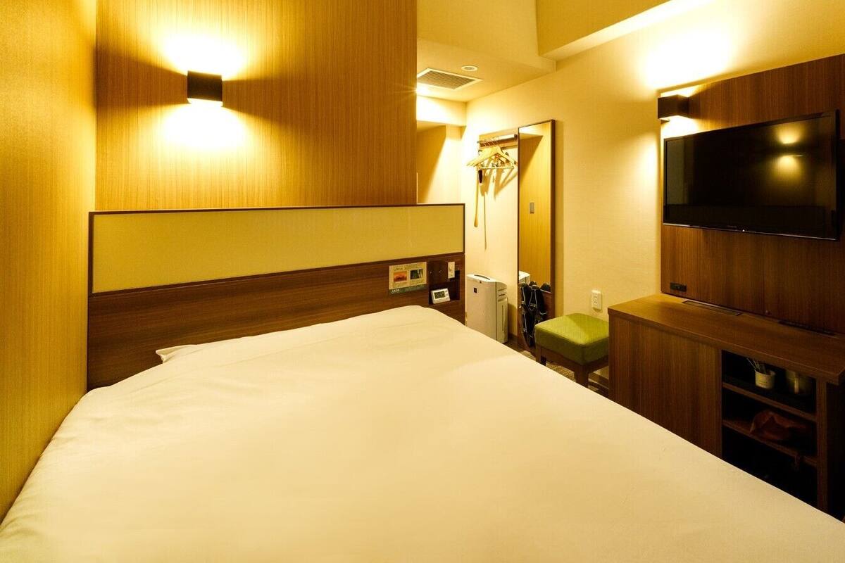 Habitación de hotel con una cama, un televisor de pantalla plana montado en la pared, un taburete verde pequeño y un mueble de madera debajo del televisor.