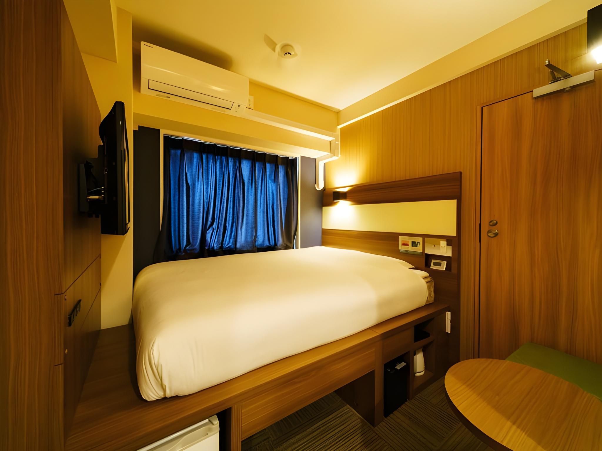 Una habitación compacta y bien iluminada con una cama, un televisor, una mesita y un armario.