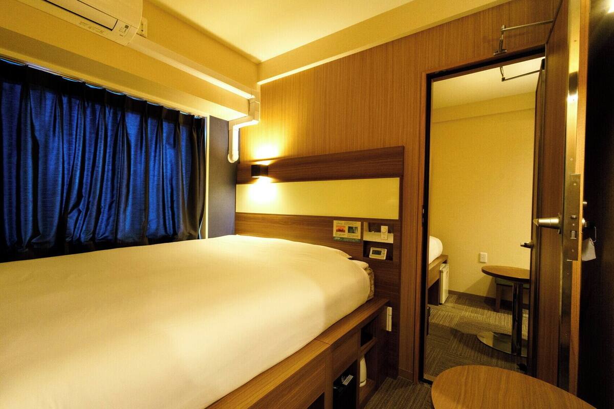 Una habitación de hotel con cama, mesita de noche, lámpara, espejo, puerta y una pequeña mesa.