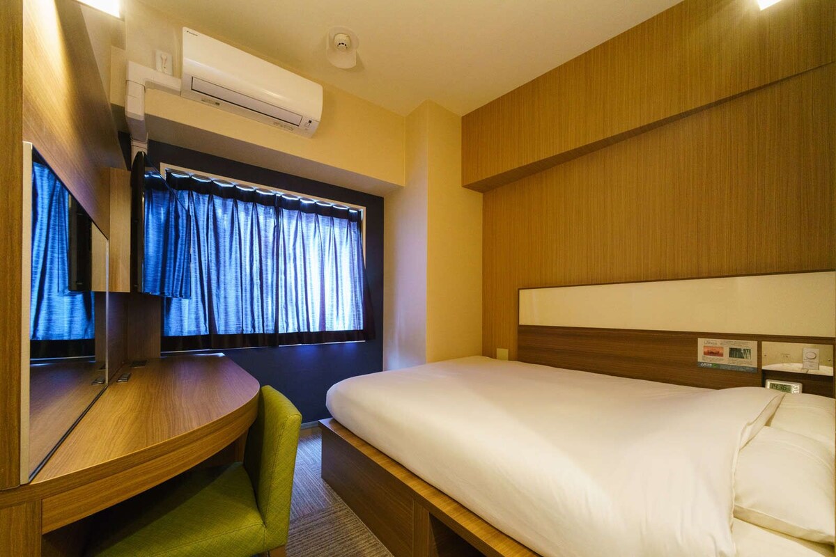 Ein Hotelzimmer mit Bett, Schreibtisch, Stuhl, einem Fenster mit Vorhängen und einer Klimaanlage.