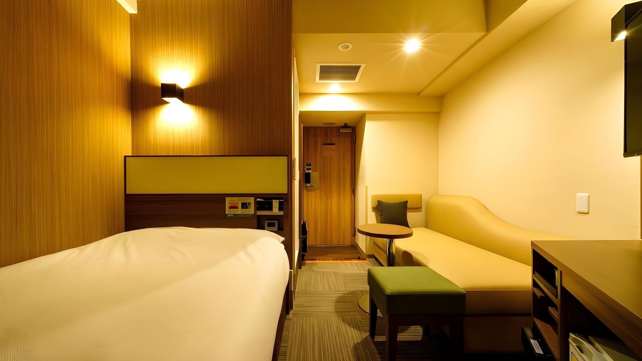 A hotel room with a bed, a sofa, a small table, a TV, and a minibar.