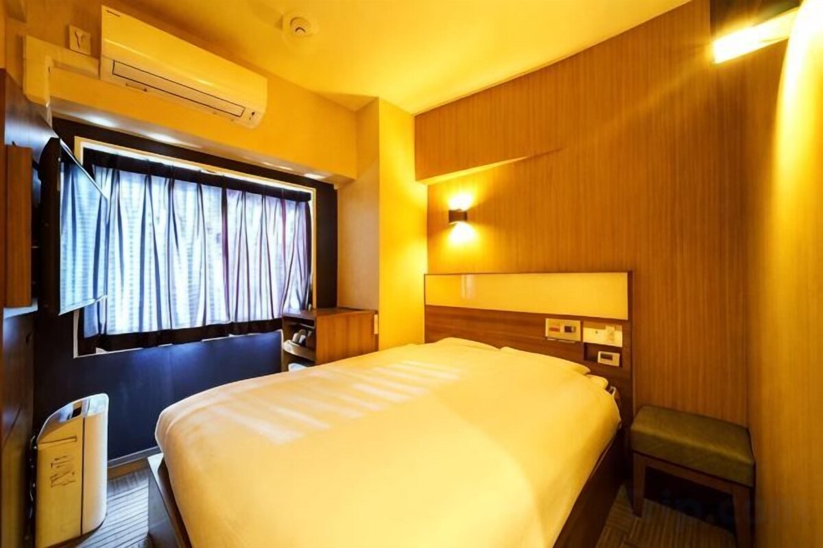 A hotel room with a large bed, bedside tables, a window with curtains, an air conditioning unit, and a wall-mounted light fixture.