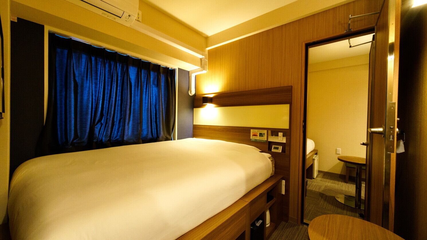 A hotel room with a bed, a bedside table, a mirror, a door, and a small table.