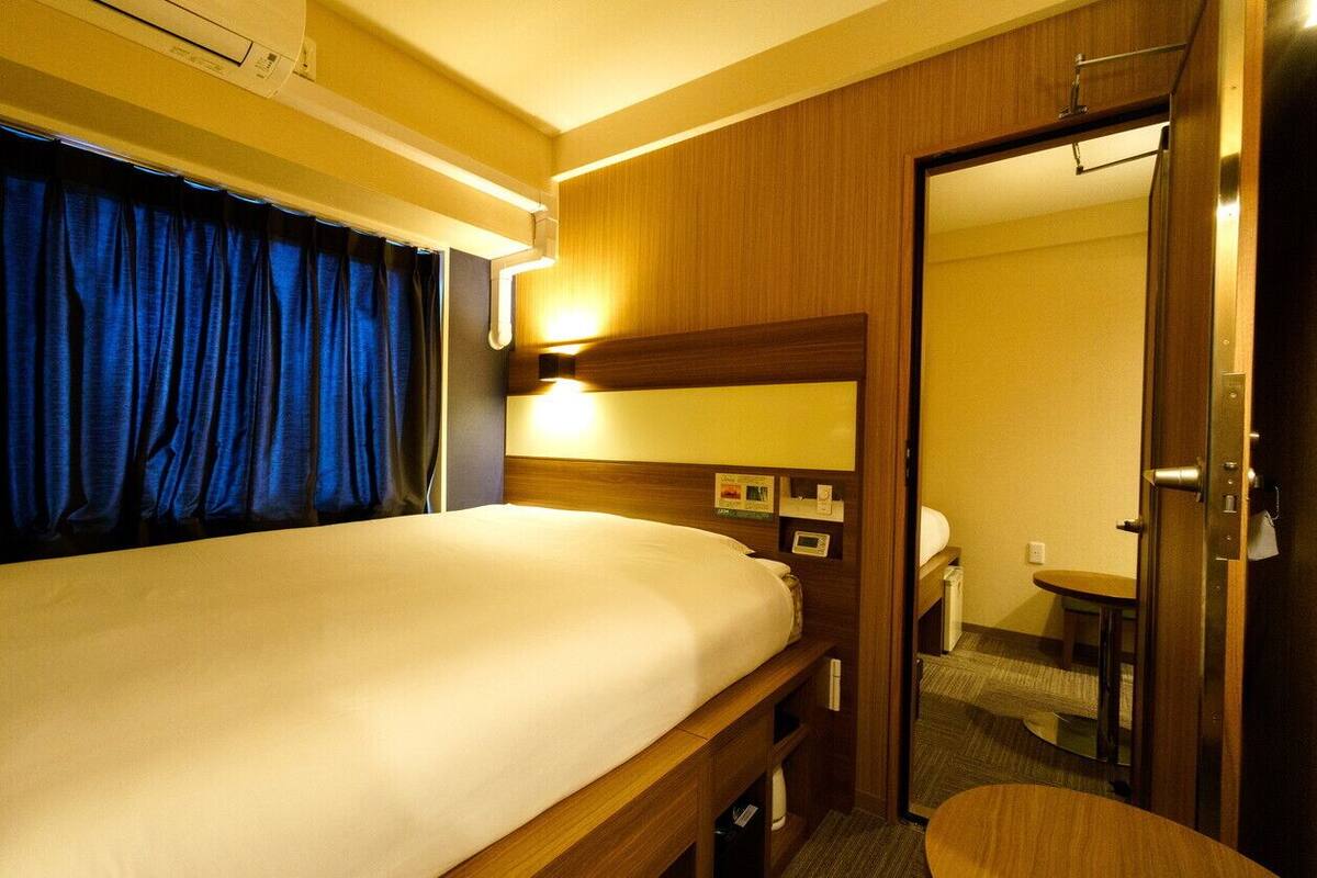 Una habitación de hotel con cama, mesita de noche, espejo, puerta y una pequeña mesa.