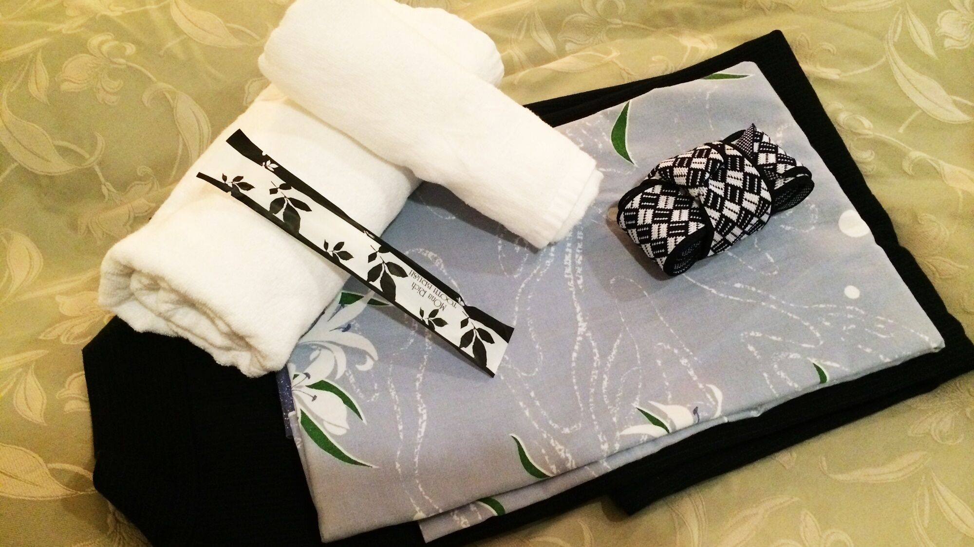 A neatly folded white towel, a black and white patterned item, and a grey pillow with floral designs on a bed with a patterned bedspread.