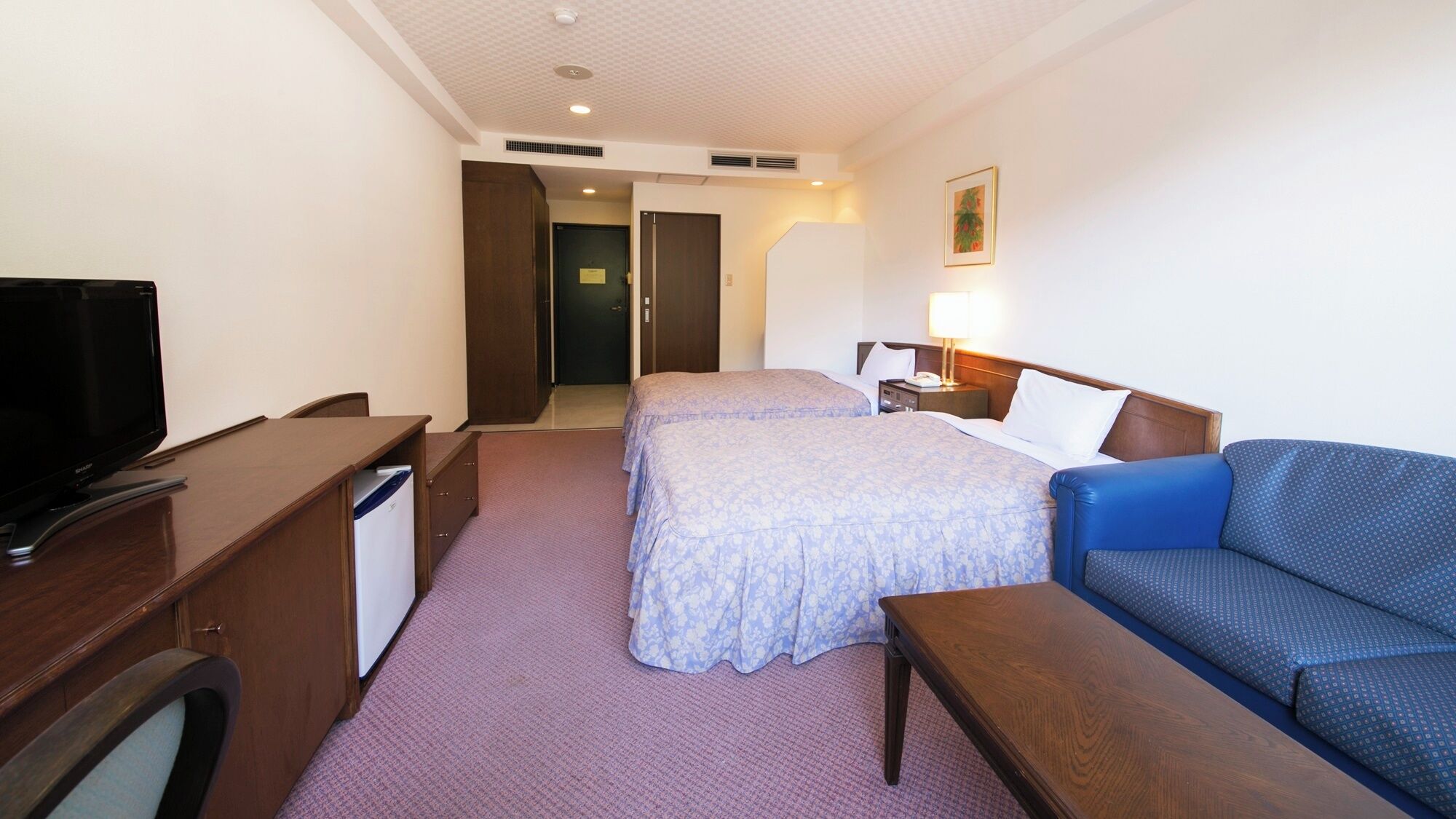 A hotel room with a TV, desk, two beds, a sofa, and a nightstand.