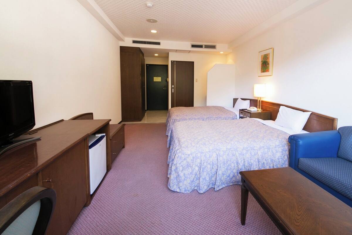 A hotel room with a TV, desk, two beds, a sofa, and a nightstand.