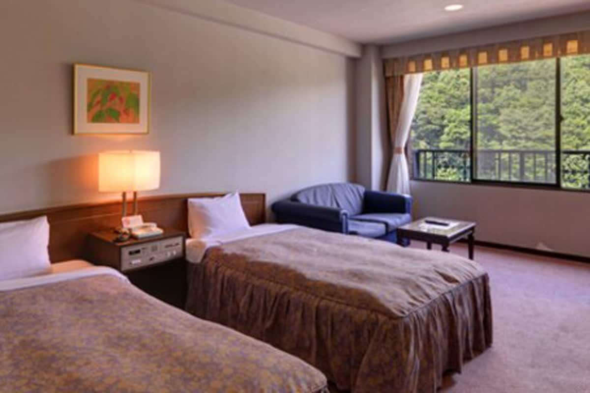 A hotel room with two beds, a desk, a chair, a lamp, and a window with a view of greenery.