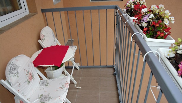 Ein Balkon mit zwei weißen Stühlen, davon einer mit rotem Tischlein, und einem Geländer mit Blumenkästen.