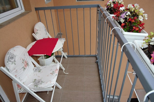 Een balkon met twee witte stoelen, waarvan er één een rode tafelkleed heeft, en een leuning met bloembakken.