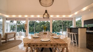 Un coin repas avec une table en bois dressée pour six personnes, entourée de chaises blanches, et offrant une vue sur un jardin luxuriant et une piscine à l’extérieur.