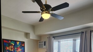 Un dormitorio con cama, ventilador de techo, ventana con cortinas, unidad de aire acondicionado y un cuadro de colores en la pared.
