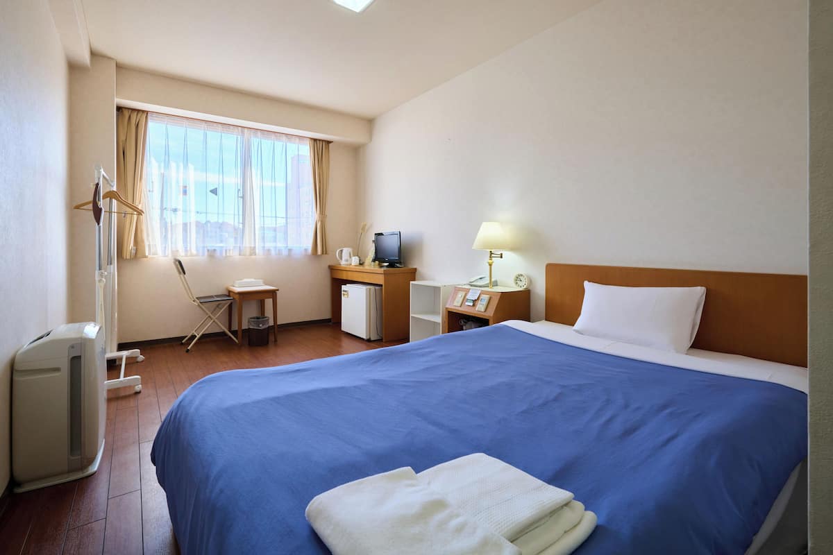 A hotel room with a large bed, a desk with a computer, a chair, and a window with curtains.