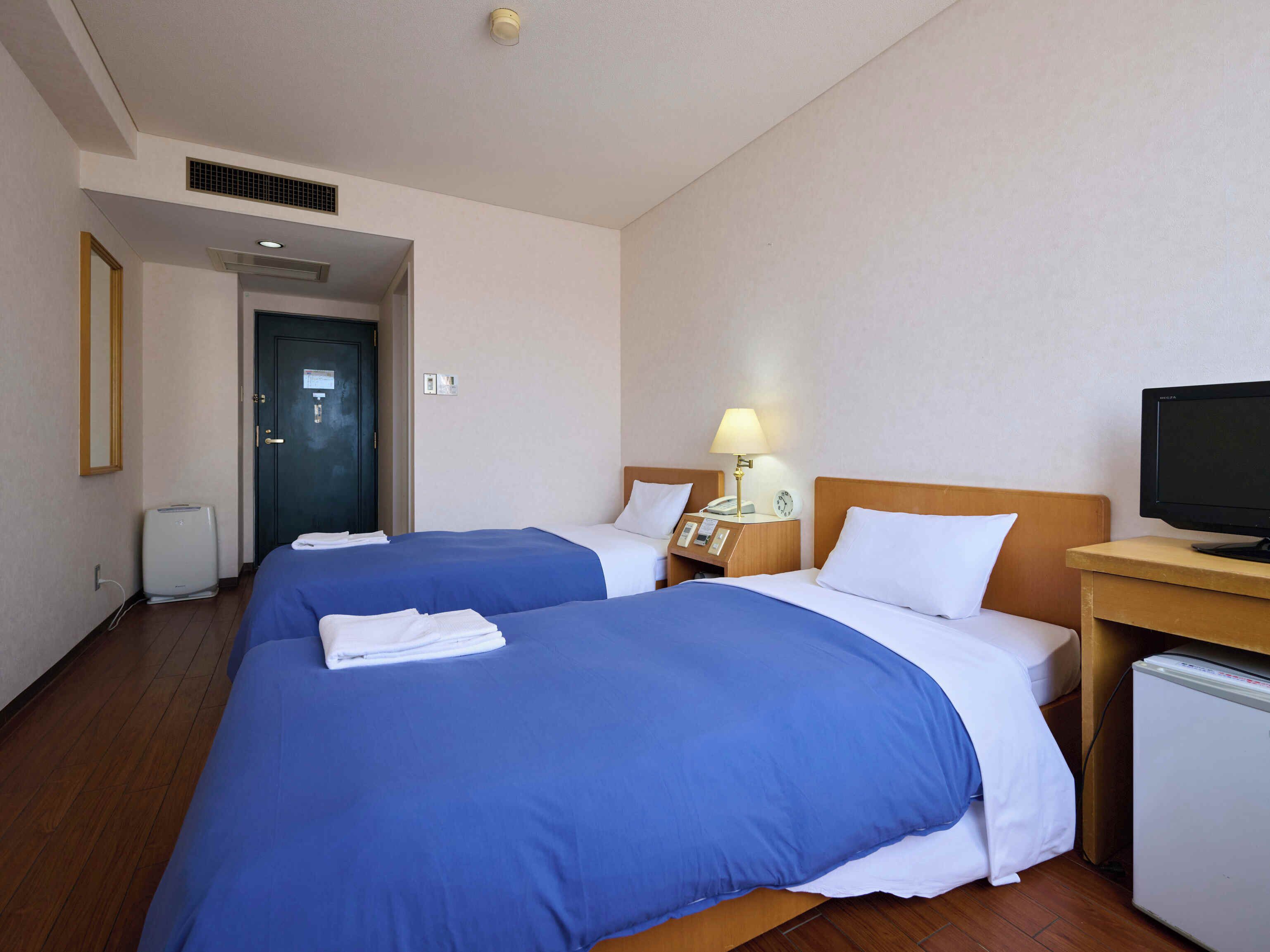 Habitación de hotel con dos camas, un escritorio con lámpara, televisión y un refrigerador pequeño.