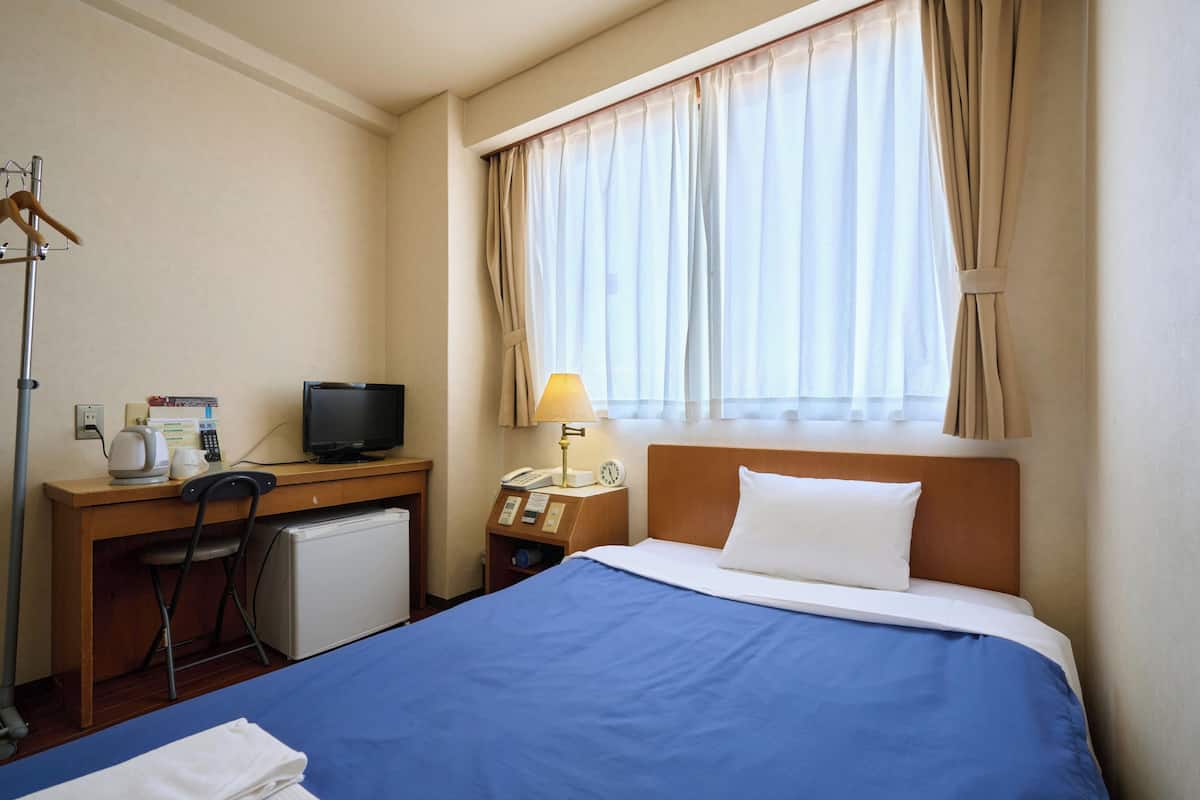 A hotel room with a bed, a desk with a computer, a chair, a lamp, and a small refrigerator.