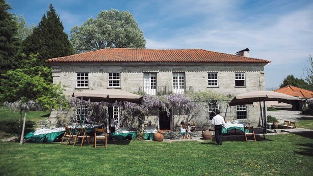 A stone building with a tiled roof, a small porch with chairs, and a garden with a table and chairs. - Casa da Arosa Rural Tourism (Gandarela)