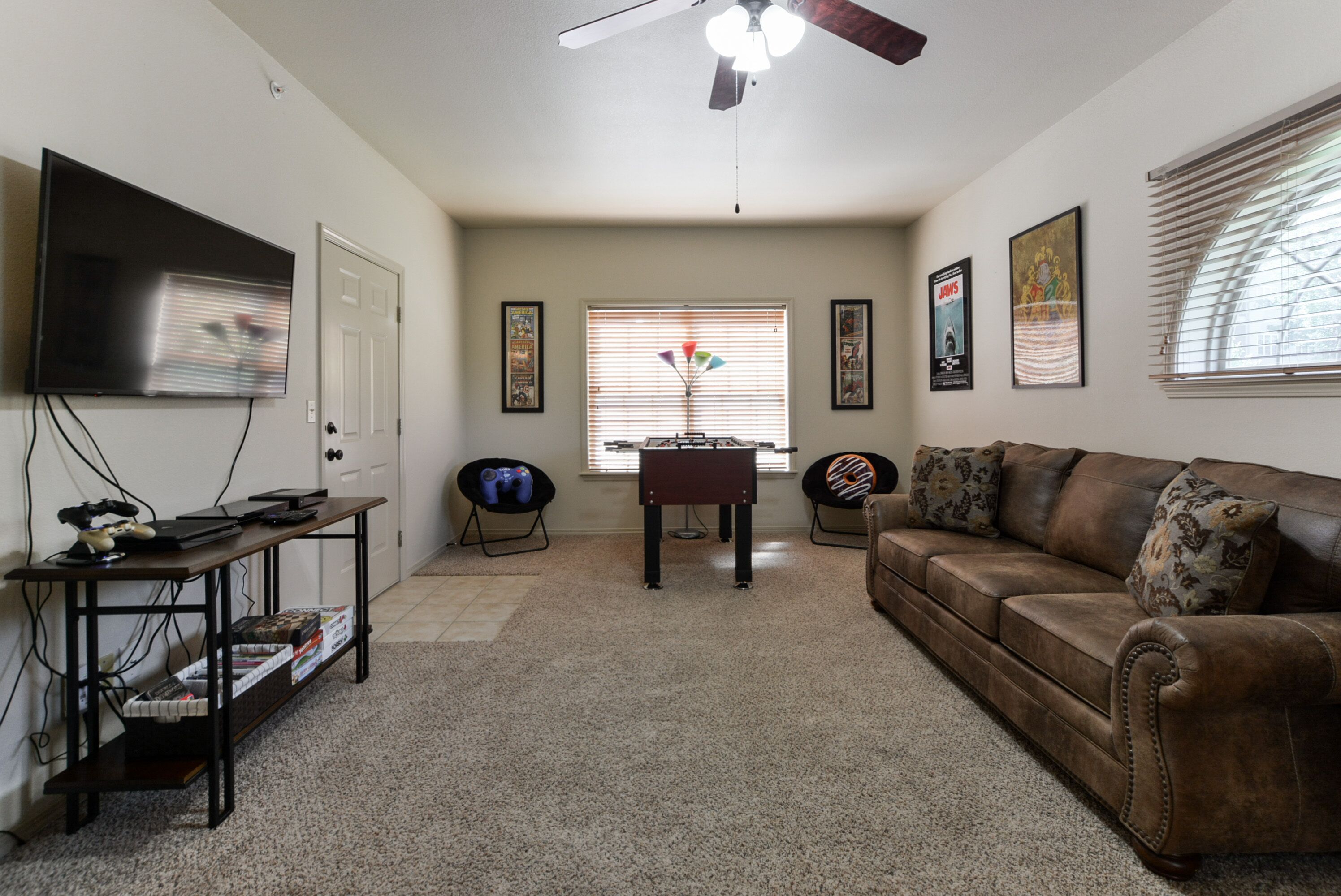 A living room with a brown leather couch, a TV mounted on the wall, a ceiling fan, and a window with blinds.