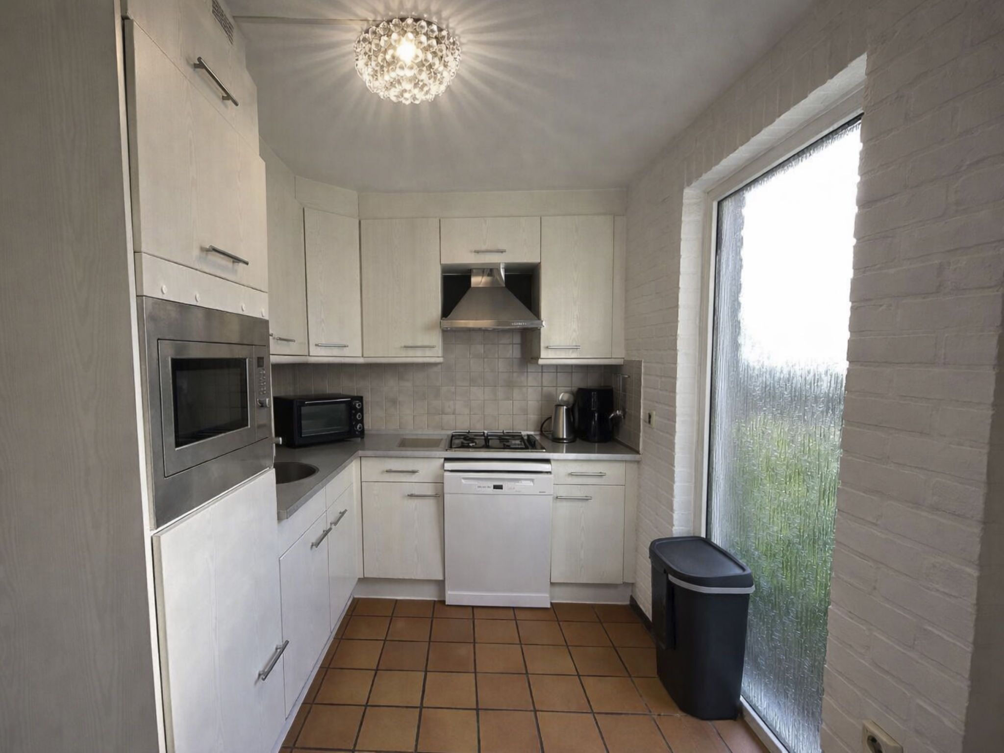 A compact kitchen with white cabinets, a built-in oven, microwave, and dishwasher. There's a window with a view of greenery outside.