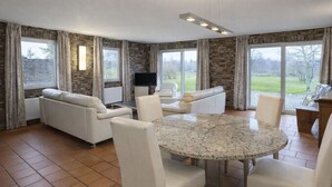A spacious living room with a round marble table, white leather sofas, and a flat-screen TV.