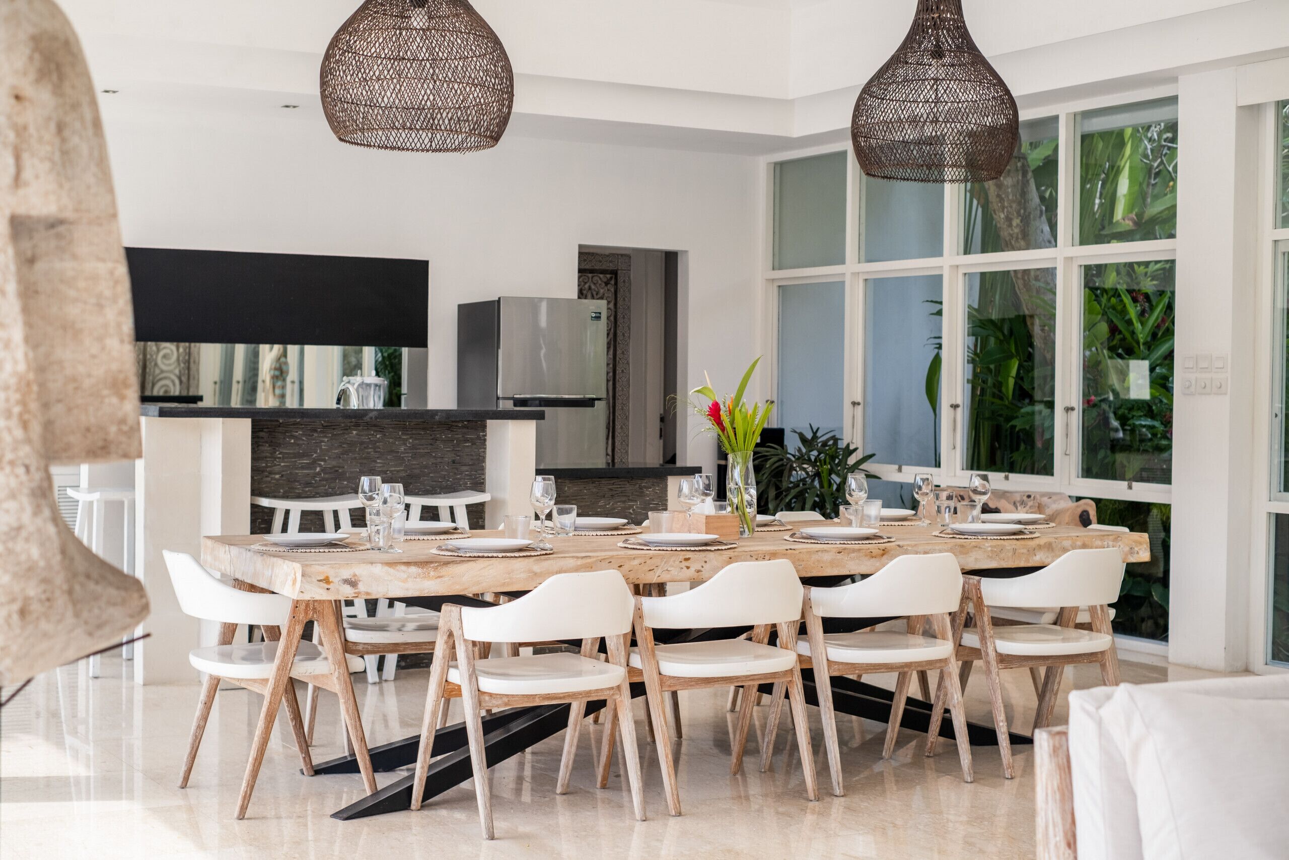 A modern dining area with a large wooden table, white chairs, and hanging wicker chandeliers.