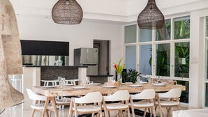 Uma sala de jantar moderna com uma grande mesa de madeira, cadeiras brancas e lustres de vime suspensos.