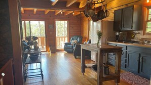 Interior acogedor de una cabaña con mesa de comedor de madera, zona de cocina con armarios y un ventanal que ofrece vistas al exterior.