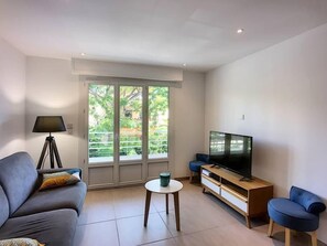 Uma sala de estar moderna com sofá, mesa de centro, televisão e uma grande janela com vista para vegetação.