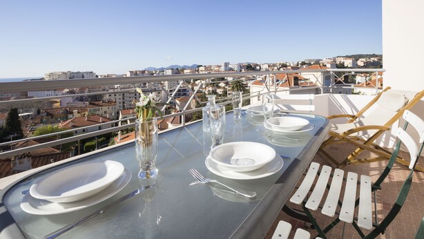 A dining table set with plates, cutlery, and glasses on a balcony overlooking a cityscape. - Bright, 63m², panoramic view of city and sea. Garage (Cannes)