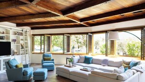 A spacious living room with a high ceiling and wooden beams, featuring a sectional sofa, blue armchairs, and a TV on a white cabinet.