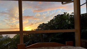 A bedroom with a large window overlooking a garden and a sunset view. - New Listing: Spacious Suite for 4x with panoramic ocean view (Kailua)