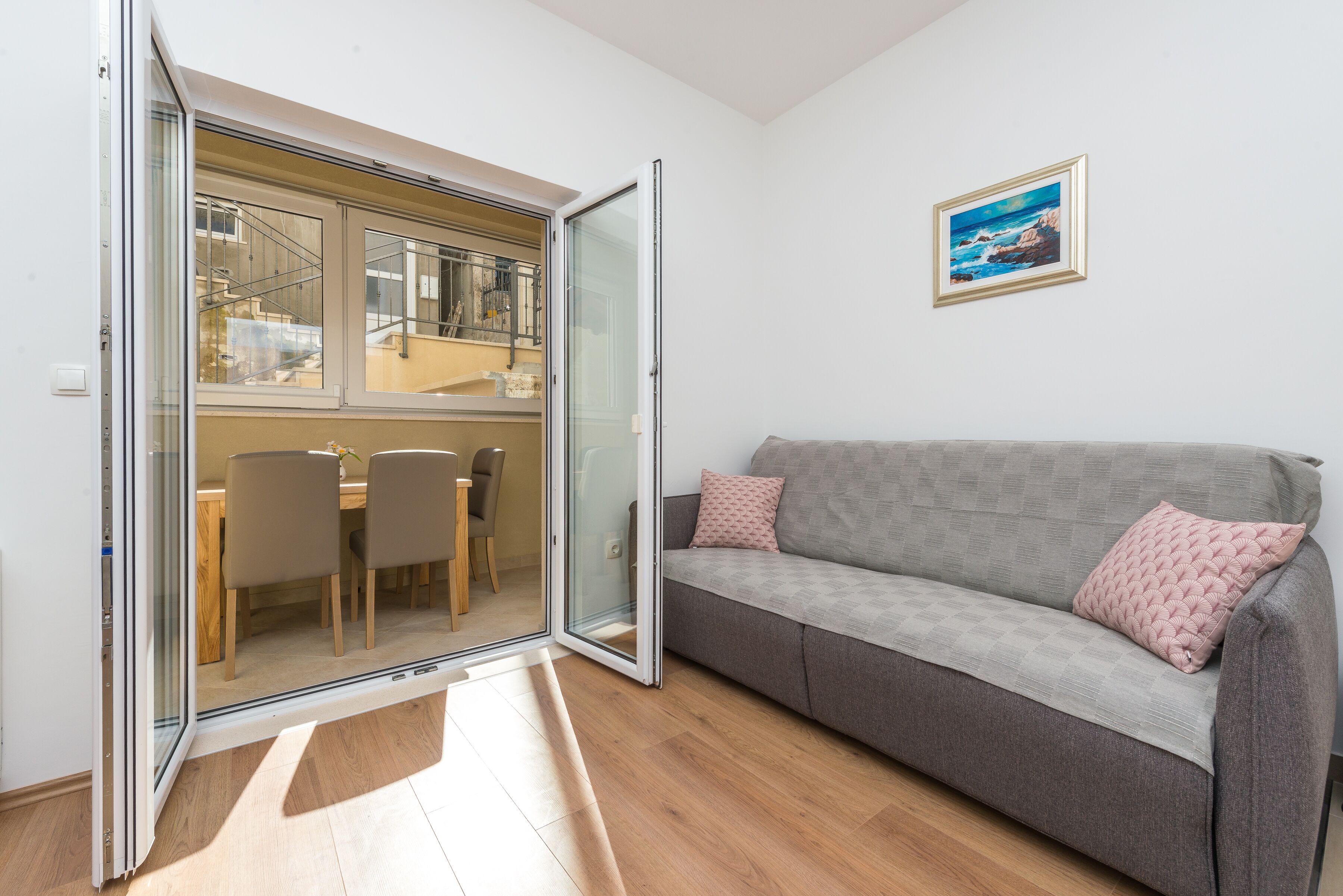 A modern living room with a grey sofa, a dining area with chairs, and a glass door leading to a balcony.