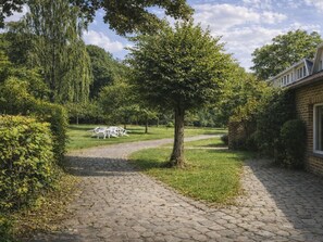 Ein gepflasterter Gartenweg führt zu einer weißen Sitzterrasse mit Tischen und Stühlen, umgeben von üppigem Grün und Bäumen.