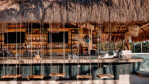 A bar area with a thatched roof, wooden bar counter, and several hanging wooden seats. - Mayan Monkey Tulum | Social Hotel (Tulum)