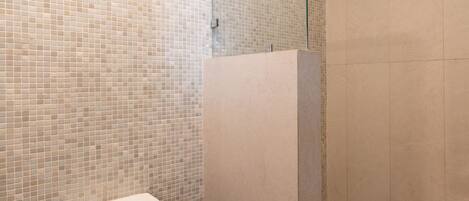 A modern bathroom with a tiled wall, a glass shower enclosure, and a wooden bench with towels.