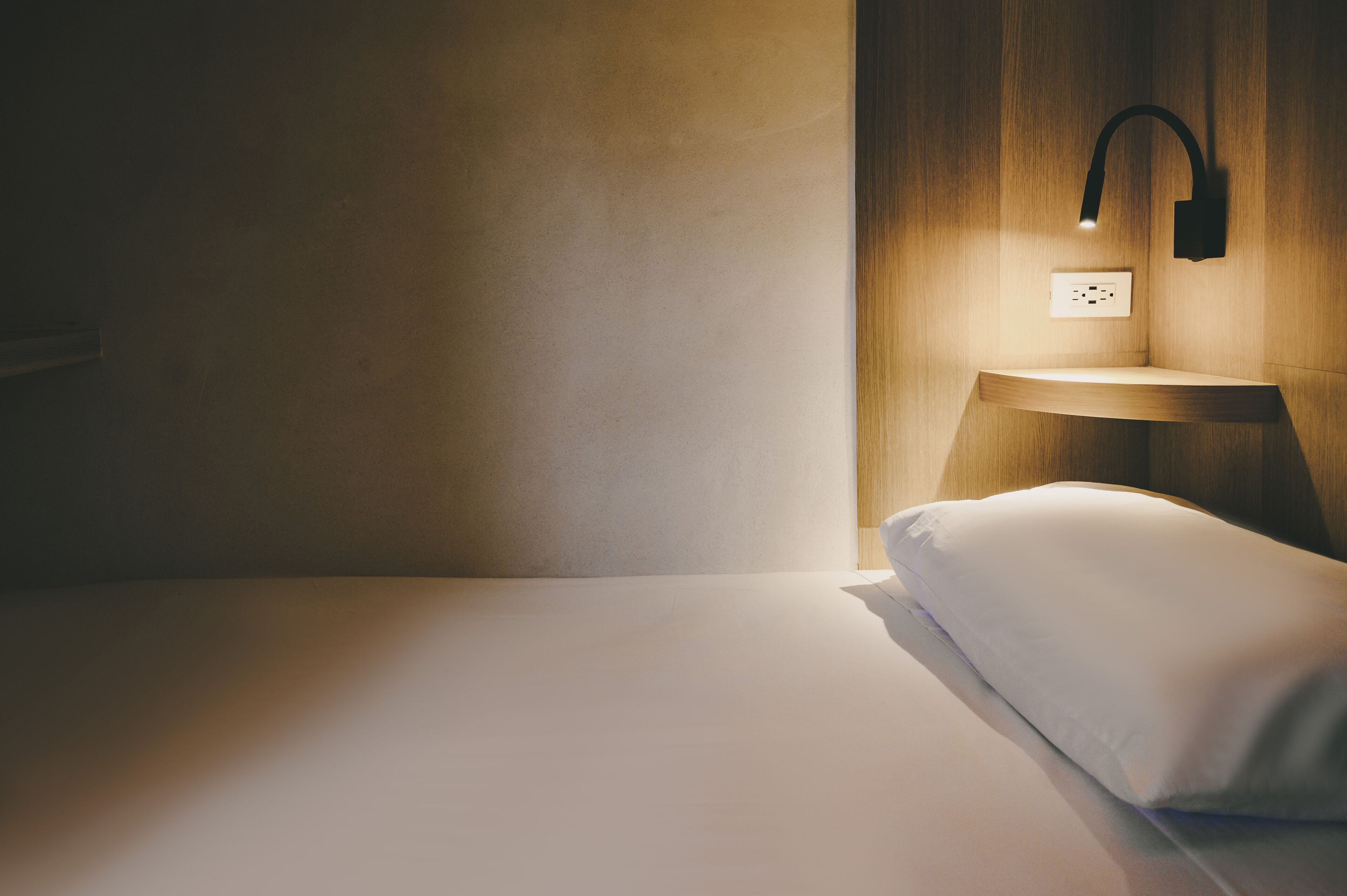 A minimalist hotel room with a single bed, a bedside table, a wall-mounted light, and a power outlet.