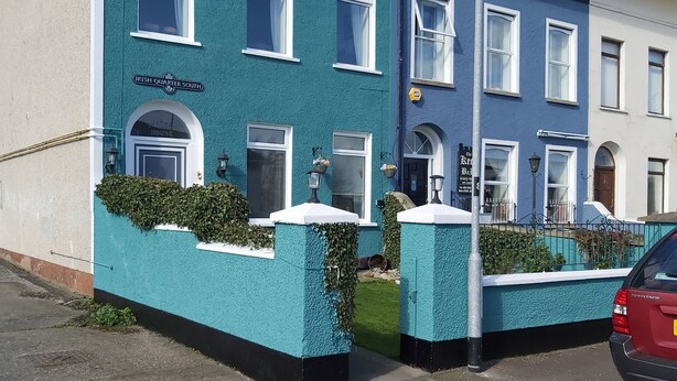 A row of two-story buildings with a teal facade, white window frames, and a sign reading "Irish Quarter South". - The Tramway House (Carrickfergus)