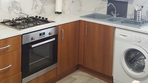 A compact kitchen with wooden cabinets, a gas stove, an oven, a washing machine, and a marble backsplash. - The Tramway House (Carrickfergus)