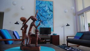 Een woonkamer met een houten sculptuur, een groot blauw abstract schilderij, een televisie en een bank met kussens.