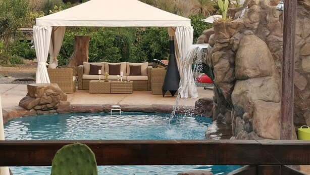 Ein Poolbereich mit Wasserfall, einer überdachten Lounge-Ecke mit Kissen und einer Kaktuspflanze im Vordergrund.