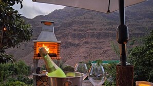 Eine Champagnerflasche, zwei Weingläser und ein Holzofen auf einem Tisch im Freien mit Blick auf die Berge.