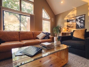 A living room with a brown leather sofa, a glass coffee table with books, a dark blue sofa, and a large window with a view of trees. - Peaceful Lakefront Retreat with Private Dock, Fire Pit, Kayaks (Gravois Mills)