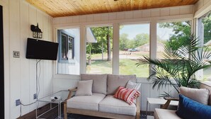 Un salon chaleureux avec un plafond en bois, un canapé agrémenté de coussins, un téléviseur fixé au mur et de grandes fenêtres offrant une vue sur l’extérieur.