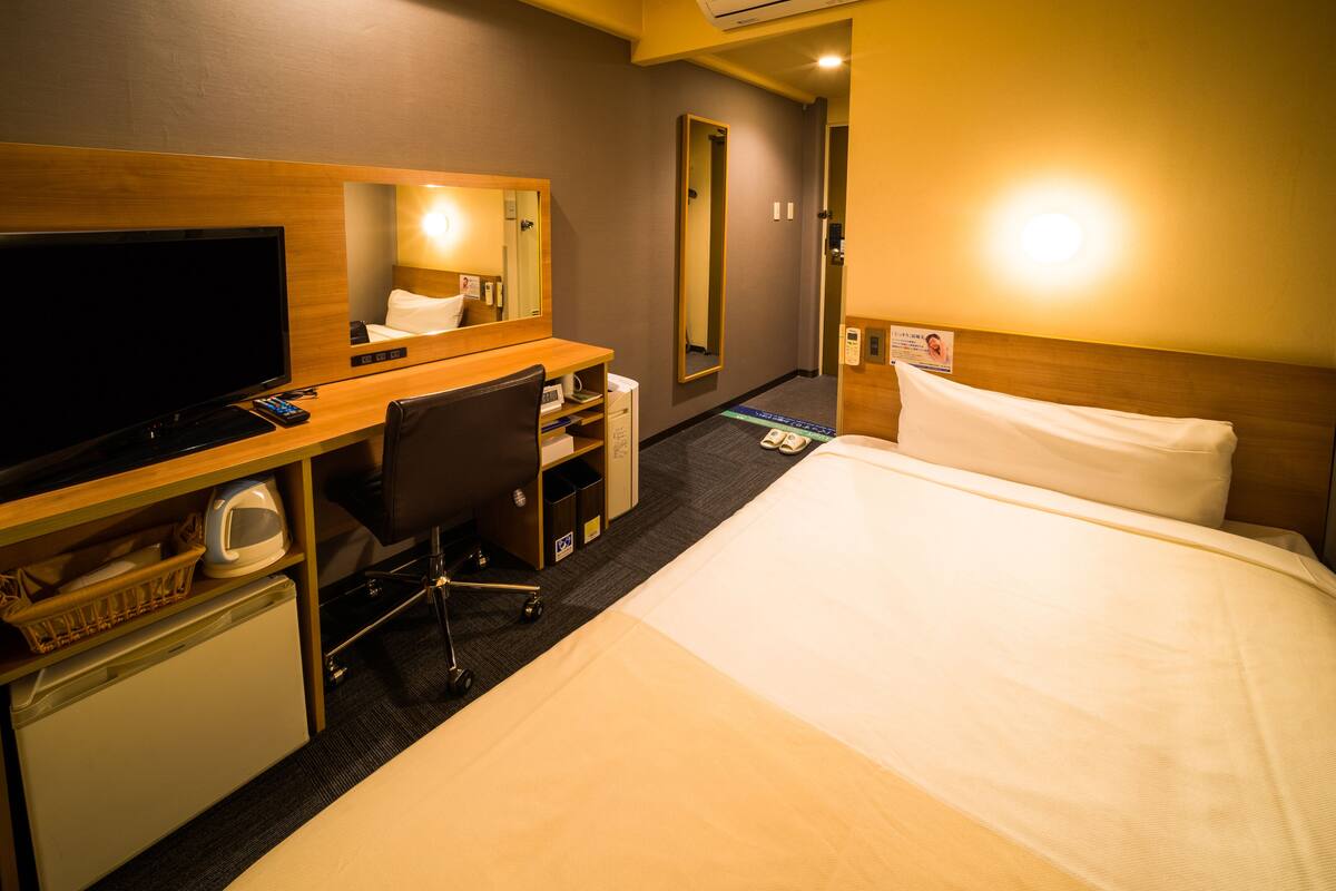 Habitación de hotel con cama, escritorio, computadora, televisor, espejo y un refrigerador pequeño.