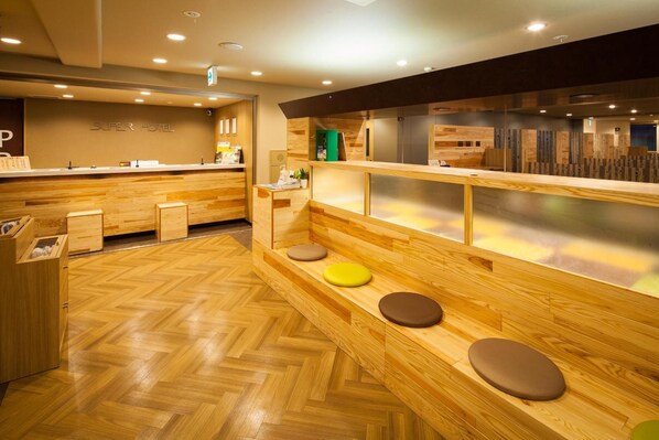 Zona de recepción de hotel con un mostrador de madera, asientos con cojines y un elevador montado en la pared.