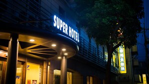 Fasaden på ett hotell med en tydlig skylt som visar texten ”Super Hotel”, en modern arkitektonisk design och ett träd i förgrunden.
