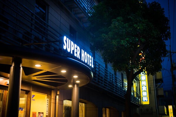 Exterior de un hotel con un llamativo letrero 'Super Hotel', diseño arquitectónico moderno y un árbol en primer plano.