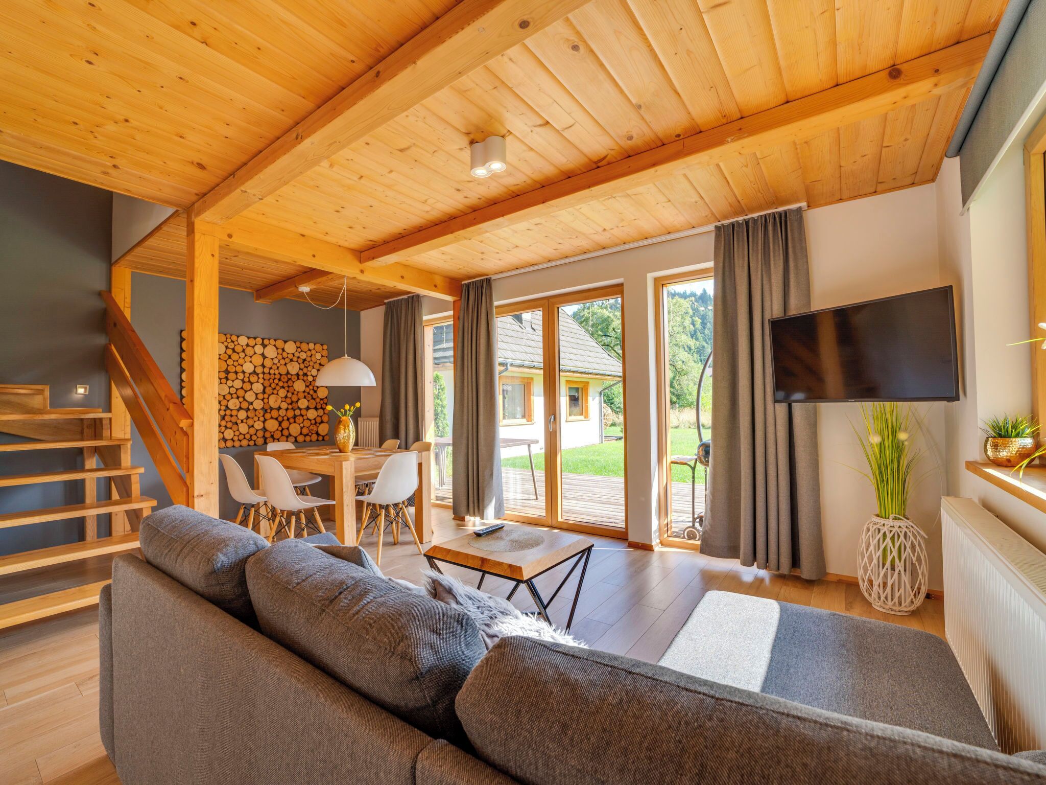 A spacious living area with a wooden ceiling, a large sofa, a dining table with chairs, and a TV mounted on the wall.