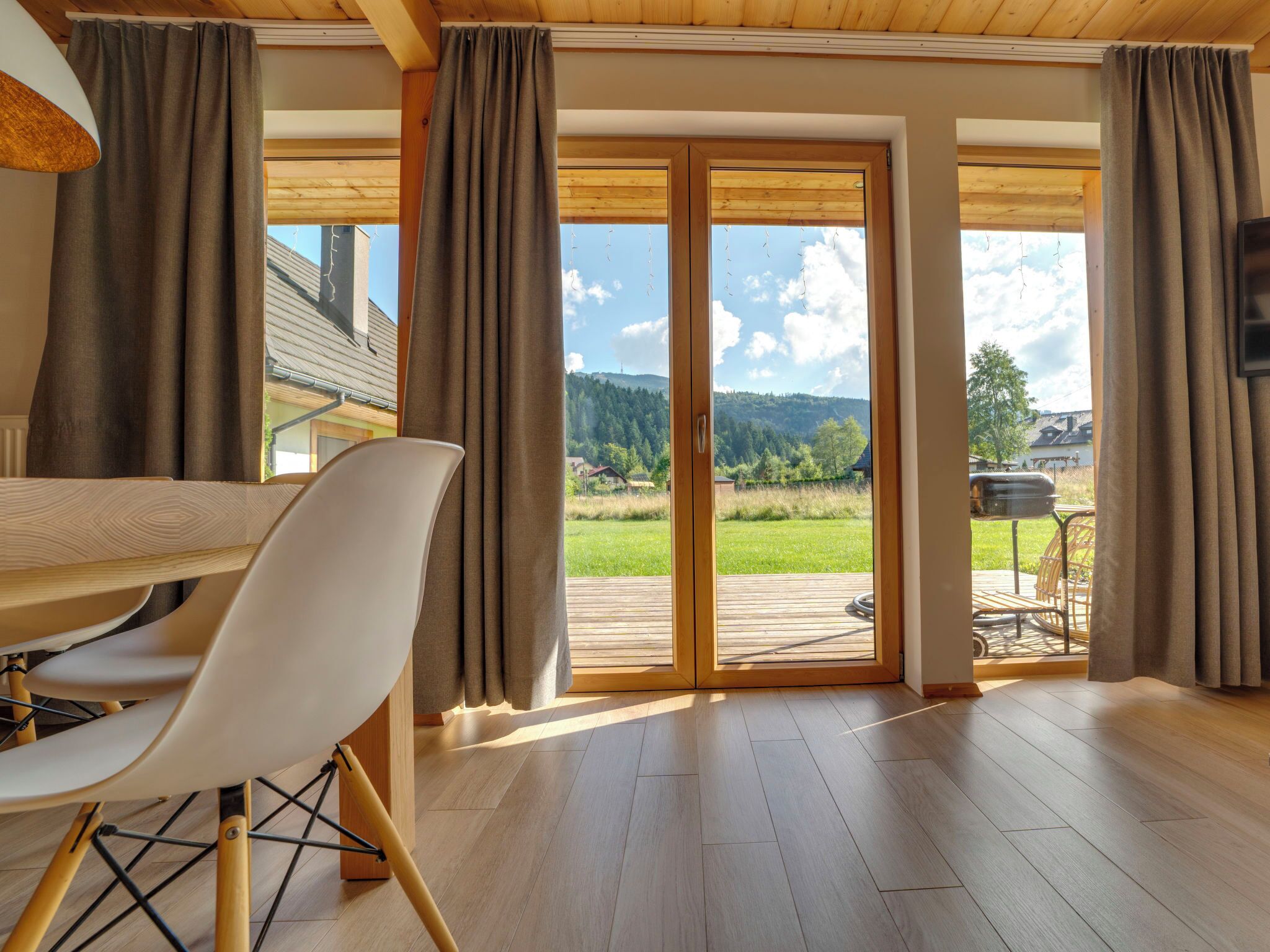 A spacious room with a wooden floor, a dining table with chairs, large sliding glass doors leading to a deck, and a view of a green landscape.
