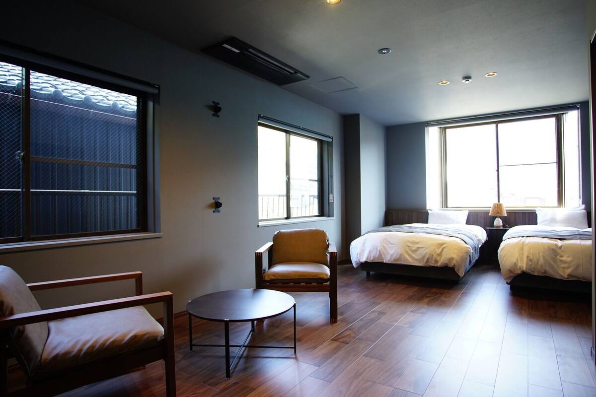 Camera d'albergo con due letti, un tavolino, una sedia e ampie finestre.